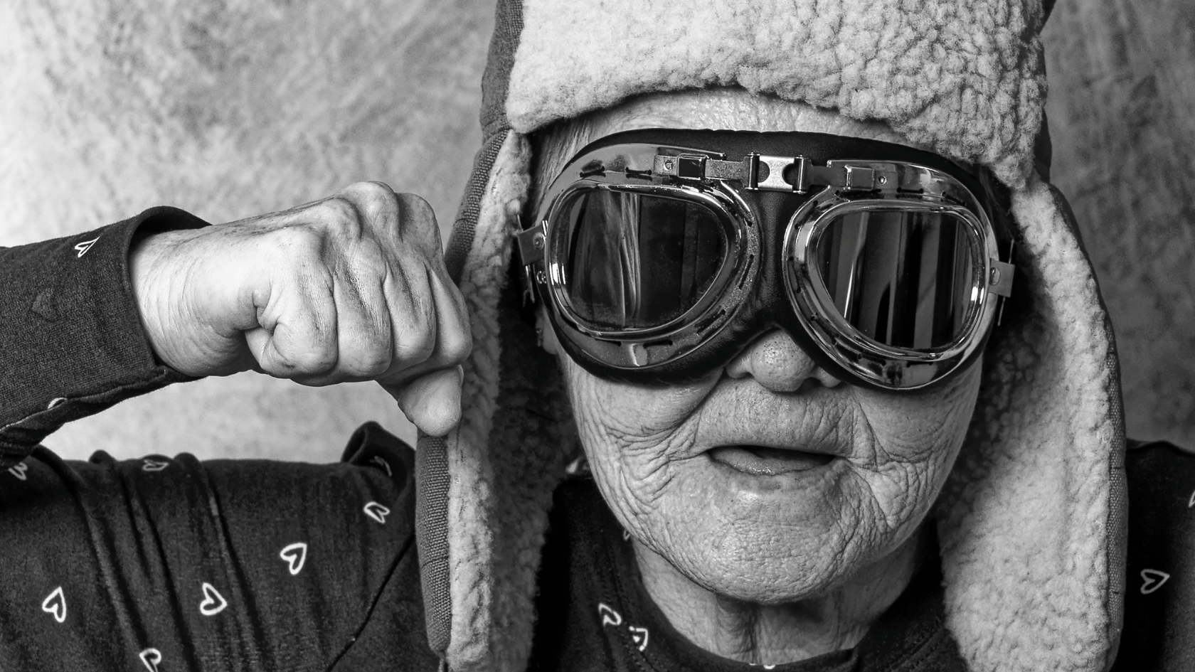 Schwarz-Weiß-Foto einer älteren Person mit Fliegerbrille und Mütze, die ihre Faust in die Kamera reckt